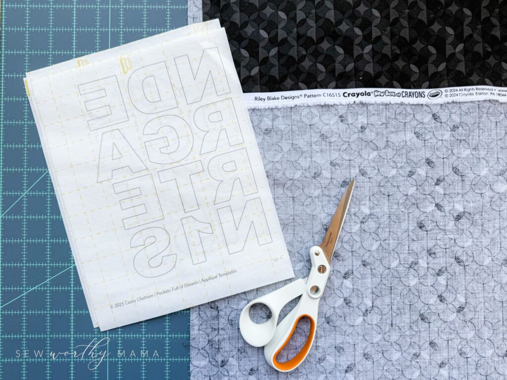 Printed appliqués next to scissors and fabric from the Crayola collection "A New Box of Crayons" for Riley Blake on top of a gridded rotary cutting mat.