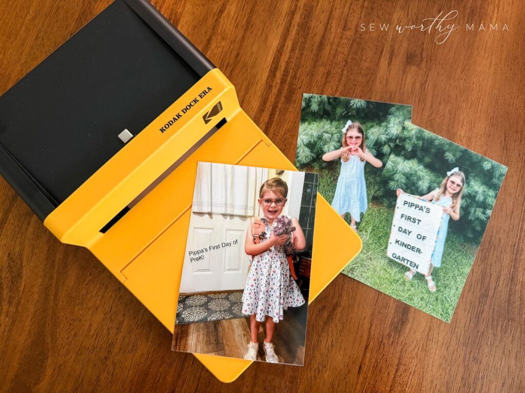 A Kodak Dock Era photo printer along with 3 printed photos. One is a child on her first day of PreK. One is the same child on her first day of kindergarten, holding the Pockets Full of Dreams sign. One is the same child making a heart with her hands.