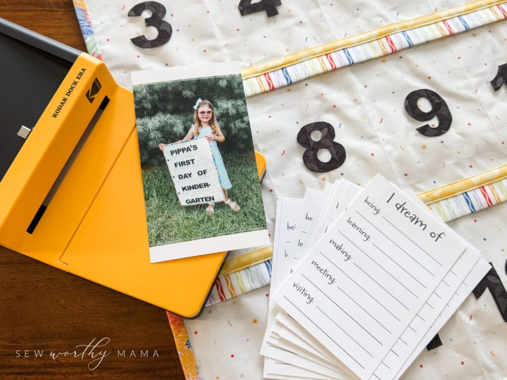 The back of a Pockets Full of Dreams project showing the pockets labeled "3, 4, 8, and 9." A Kodak Dock Era photo printer. A printed photo of a child holding her sign that says "Pippa's First Day of Kindergarten." A stack of Dream Cards that say "I dream of being...learning...making...meeting...visiting."