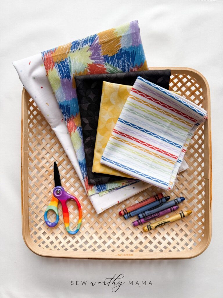 A collection of several fabrics from the A New Box of Crayons fabric collection from Riley Blake and Crayola. Fabrics are placed in a square basket along with a pair of rainbow colored scissors and a few Crayola crayons. The Sew Worthy Mama logo is present.