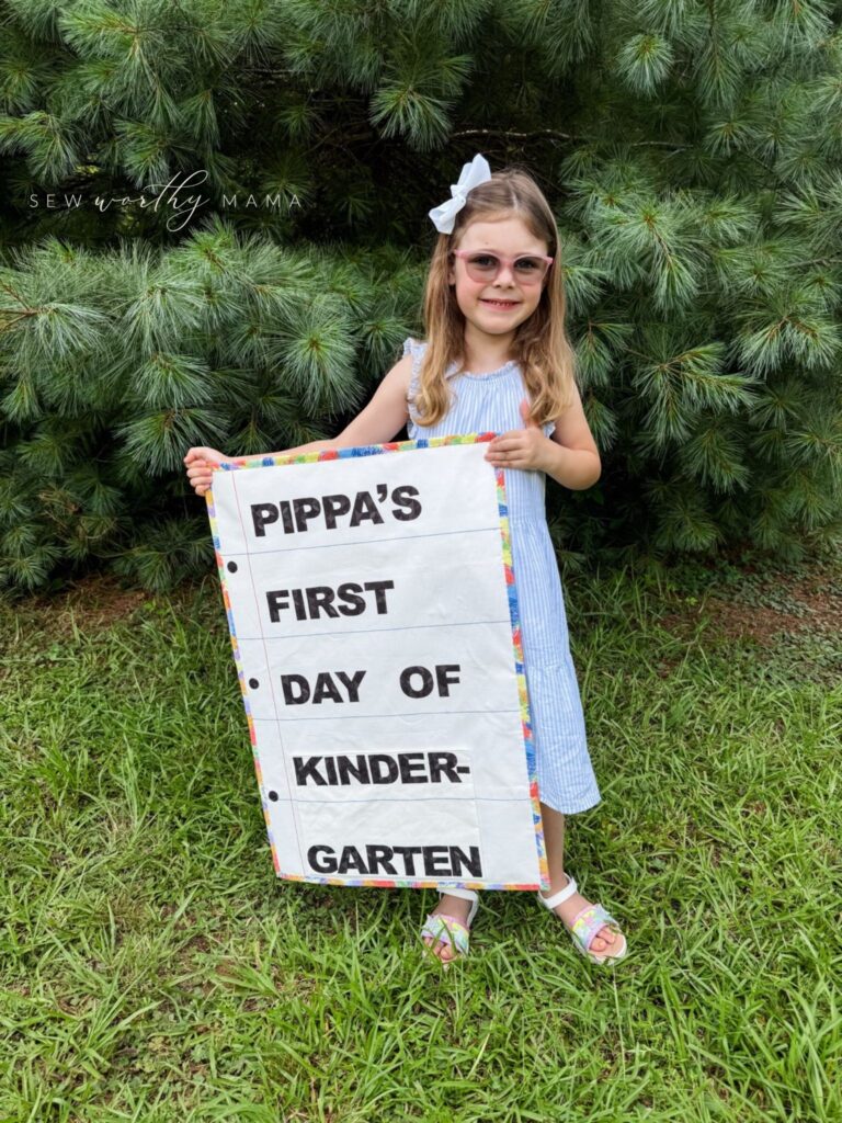 Child holding a first day of school photo prop sewn using the Pockets Full of Dreams sewing pattern. The prop says "Pippa's First Day of Kindergarten."