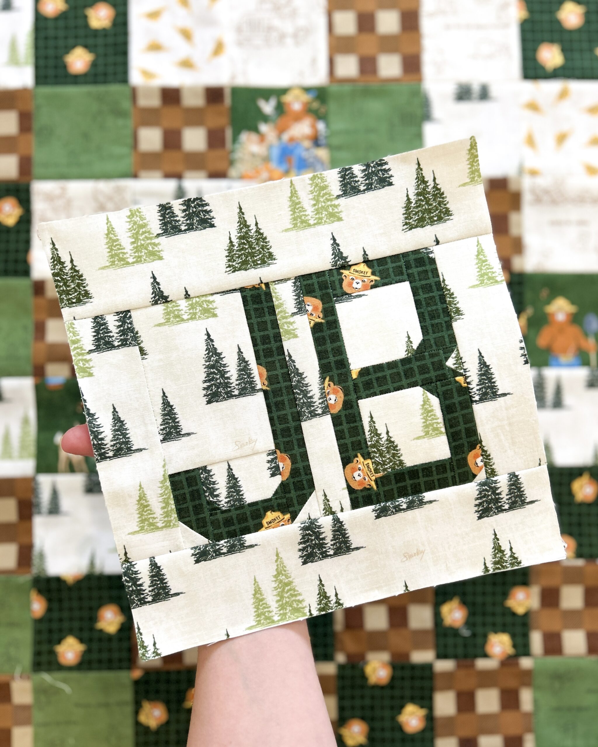 An arm holds and assembled quilt block with the initial "JB". Behind it is a completed patchwork panel.