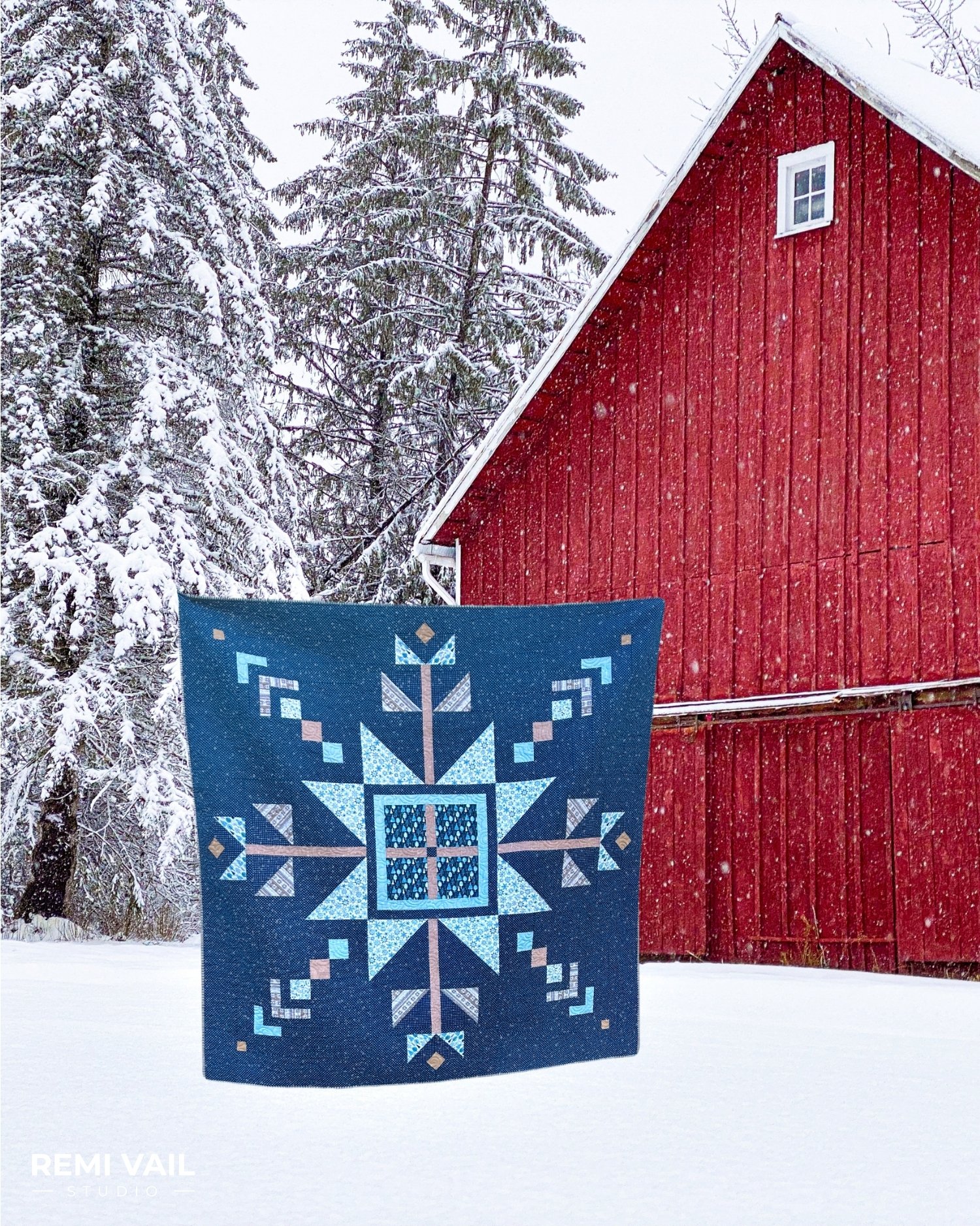 Snowflake quilt being held in the snow in front of a red barn. Lumi Quilt by Remi Vail Studio made by Tamara Darragh using the Winter Magic Fabric Collection designed by Dani Mogstad for Riley Blake Designs. 