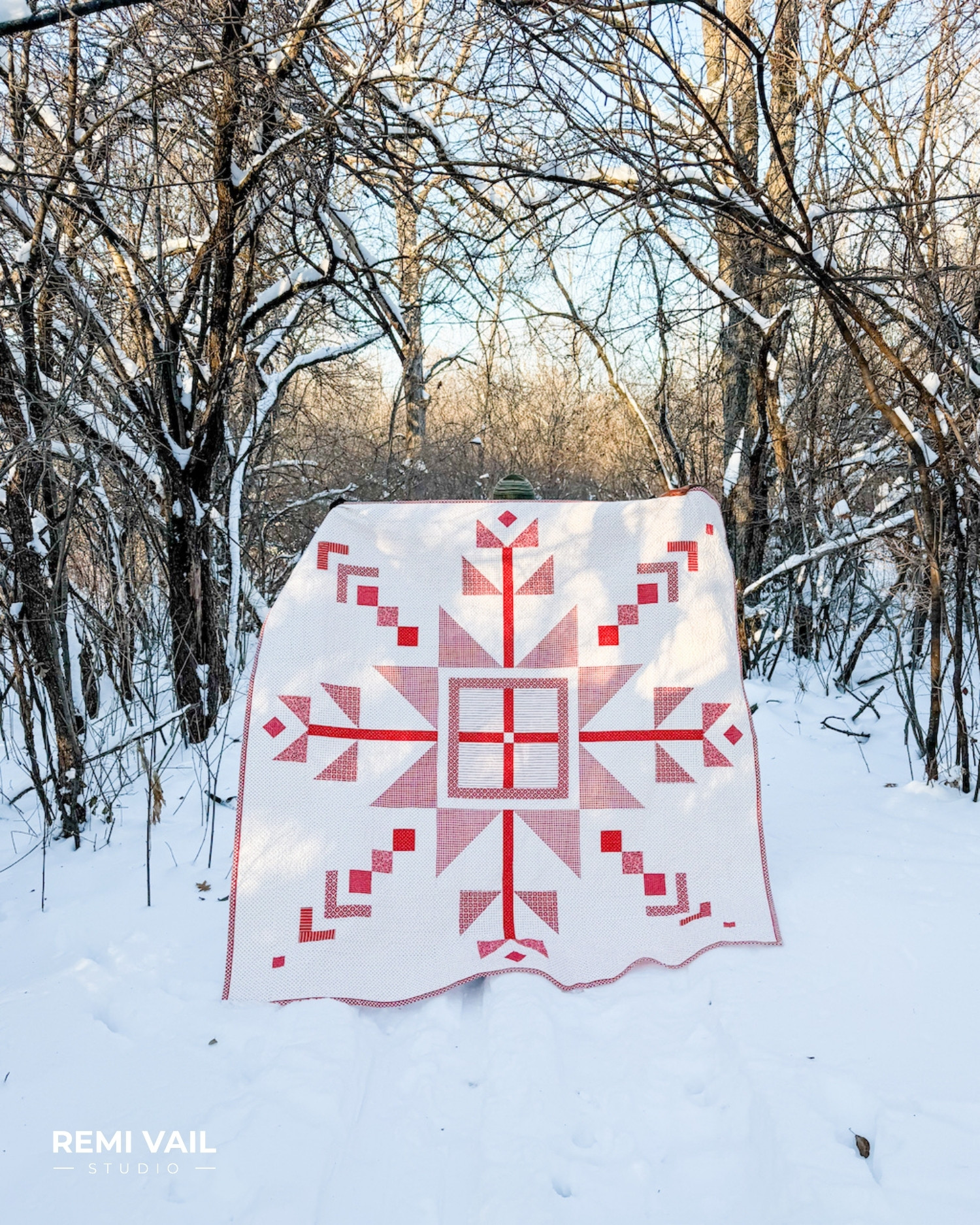 Lumi Quilt pattern by Tamara Darragh of Remi Vail Studio displayed outdoors in a snowy woodland setting, featuring a red and white snowflake medallion made with the Radiant Ruby fabric collection by Riley Blake Designs.