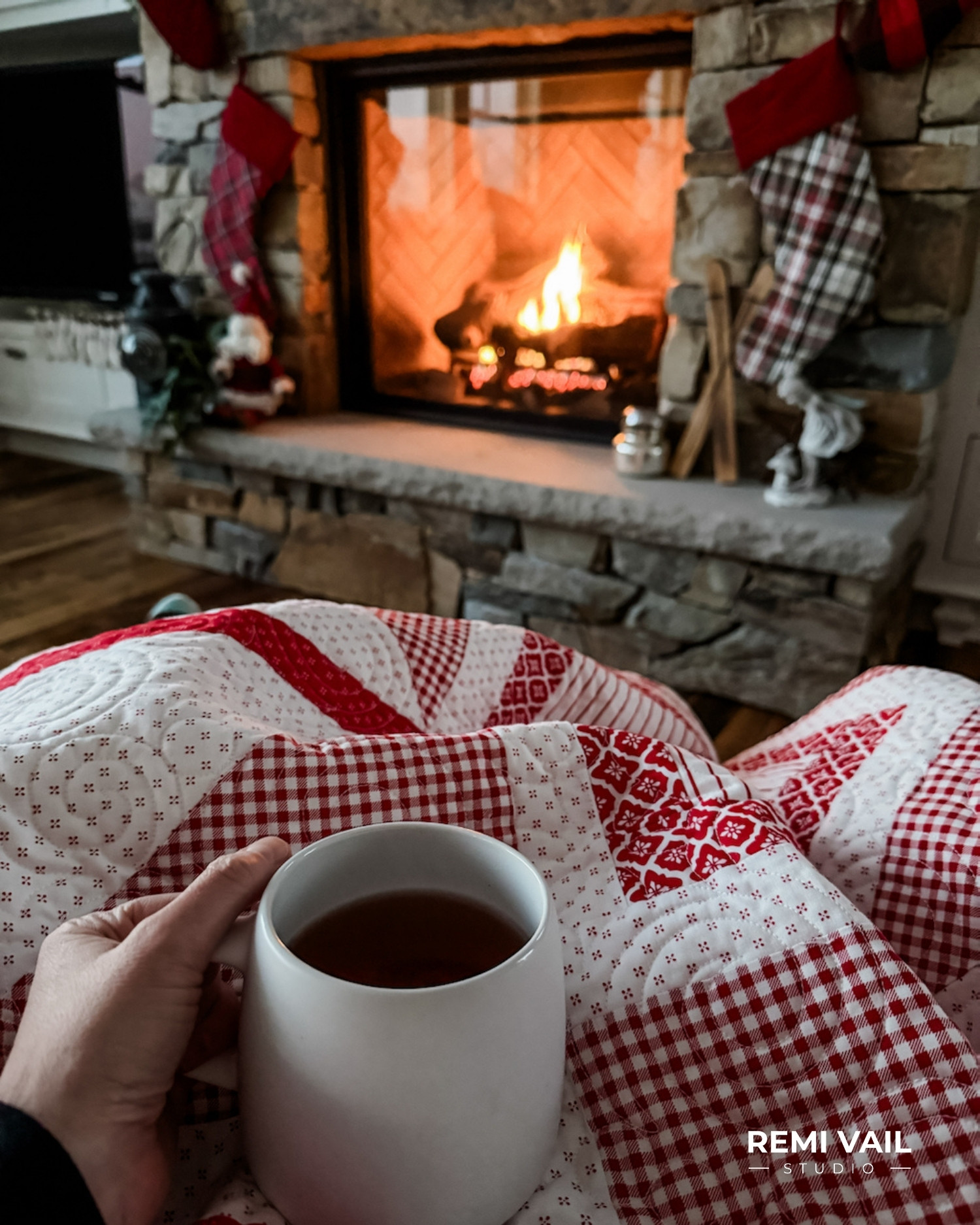 Hand holding a mug of tea while wrapped in the Lumi Quilt pattern by Tamara Darragh of Remi Vail Studio, featuring red and white fabrics from the Radiant Ruby collection by Riley Blake Designs, with a fireplace in the background.