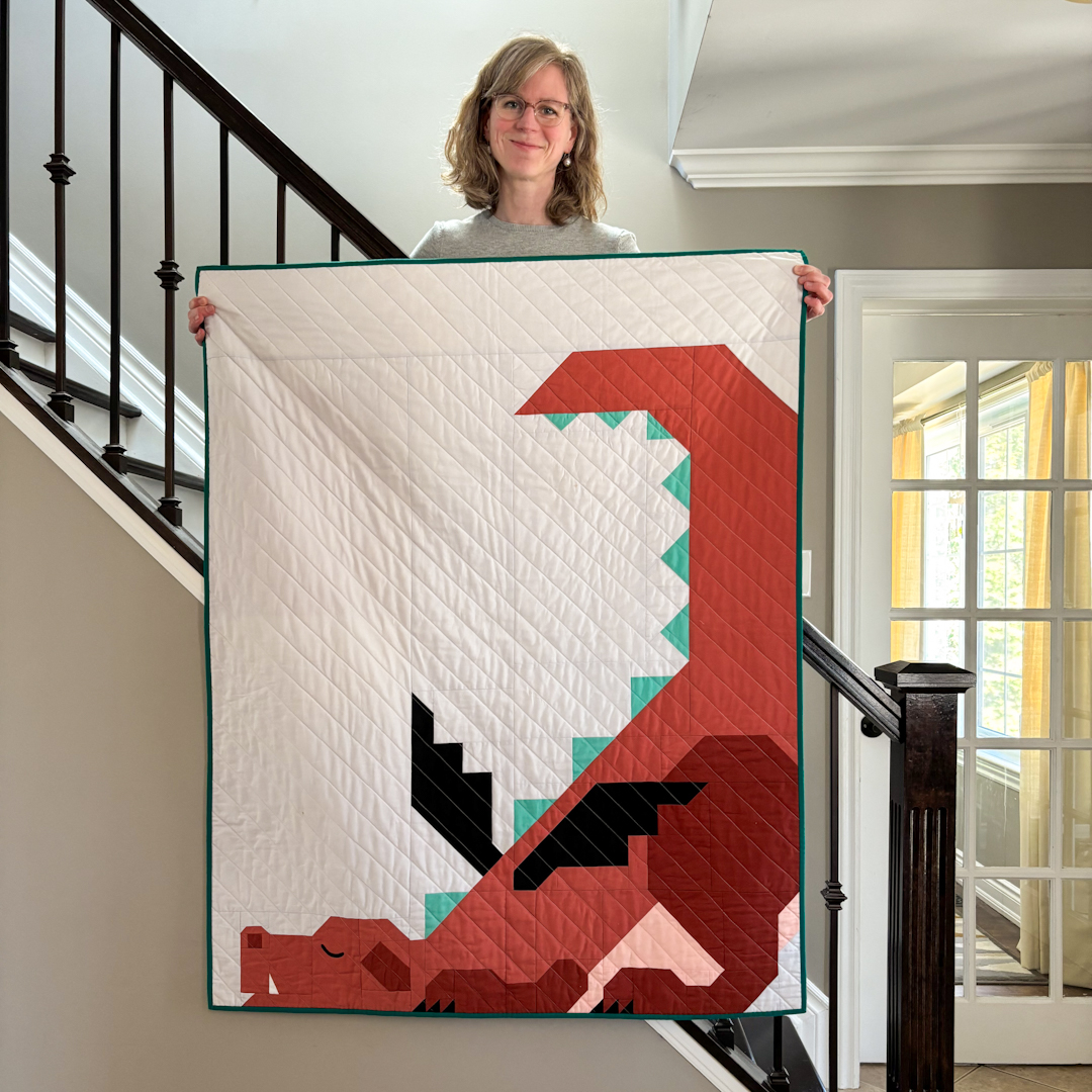 Astrid Bordush of Apples & Beavers, standing on a stairwell and showing a quilt featuring a red, sleeping dragon 