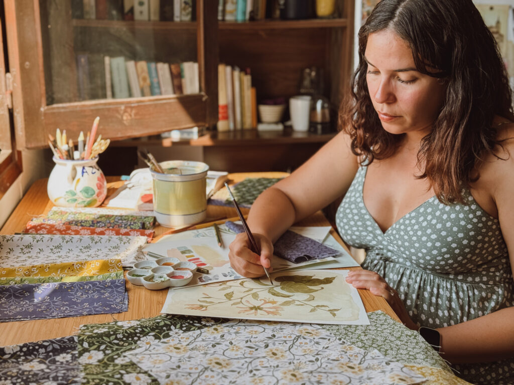 Danelys Sidron painting botanical artwork in her studio, surrounded by fabric swatches from the Before the Bloom collection for Riley Blake Designs.